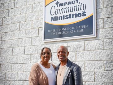 A couple stands smiling in front of Impact Community Ministries sign on a stone wall.