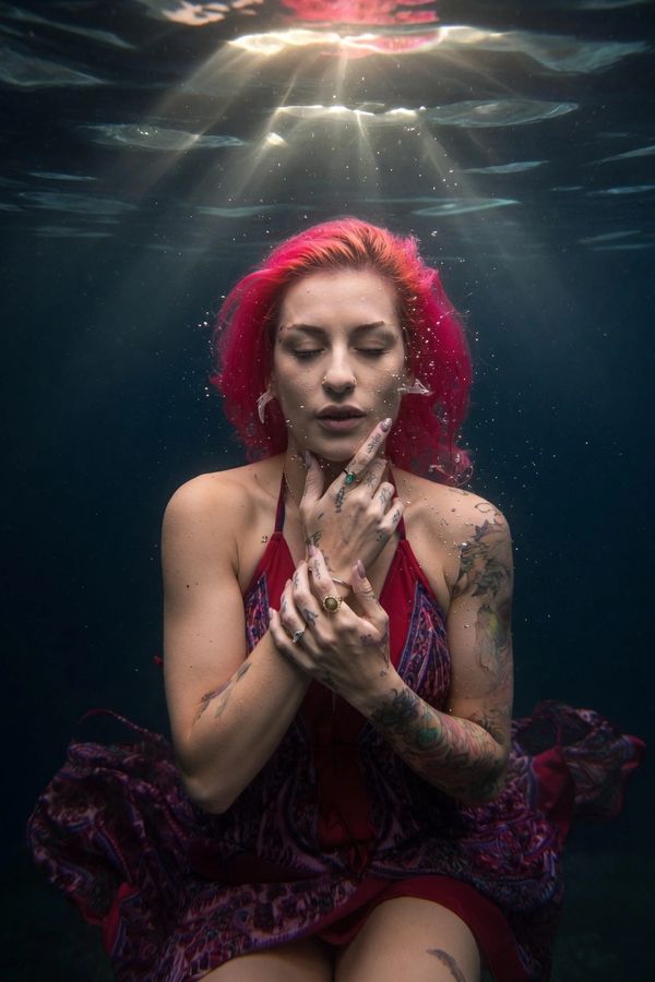 A tattooed woman with pink hair underwater, illuminated by rays of light from above.