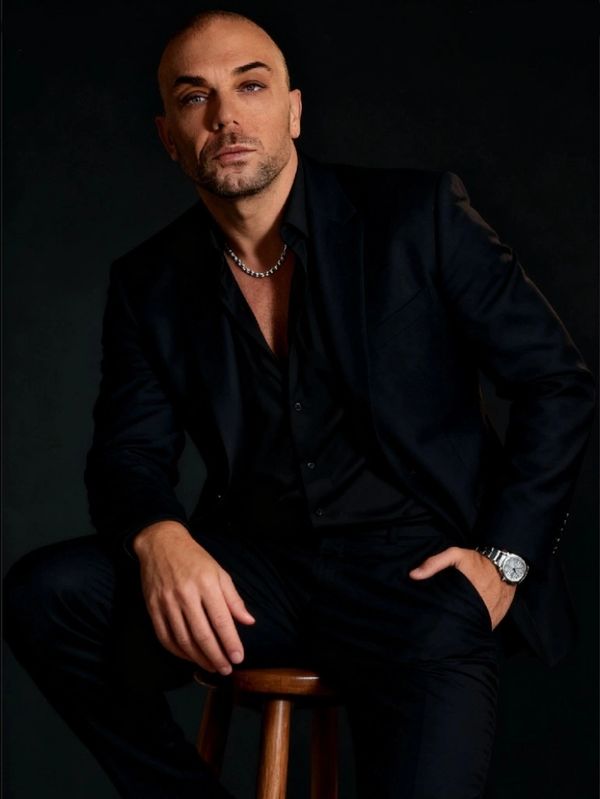 Man in black suit sitting on a wooden stool against a dark background.