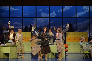 Actors perform a lively office scene on stage with vintage decor.