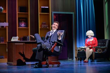 Two actors performing in a theatrical office scene, one relaxed and one serious.