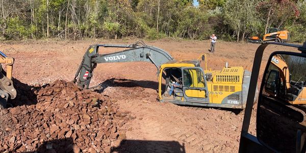 Excavator digging and moving rocks in a construction site surrounded by trees.