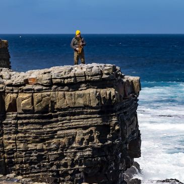 Person playing saxophone on cliff by the ocean under clear blue sky.