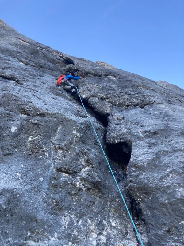 Rock climber ascending a steep cliff with safety rope under clear blue sky.