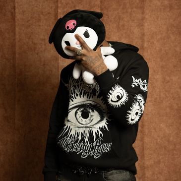 Person holding a black and white plush toy in front of their face, wearing a black hoodie with eye designs.