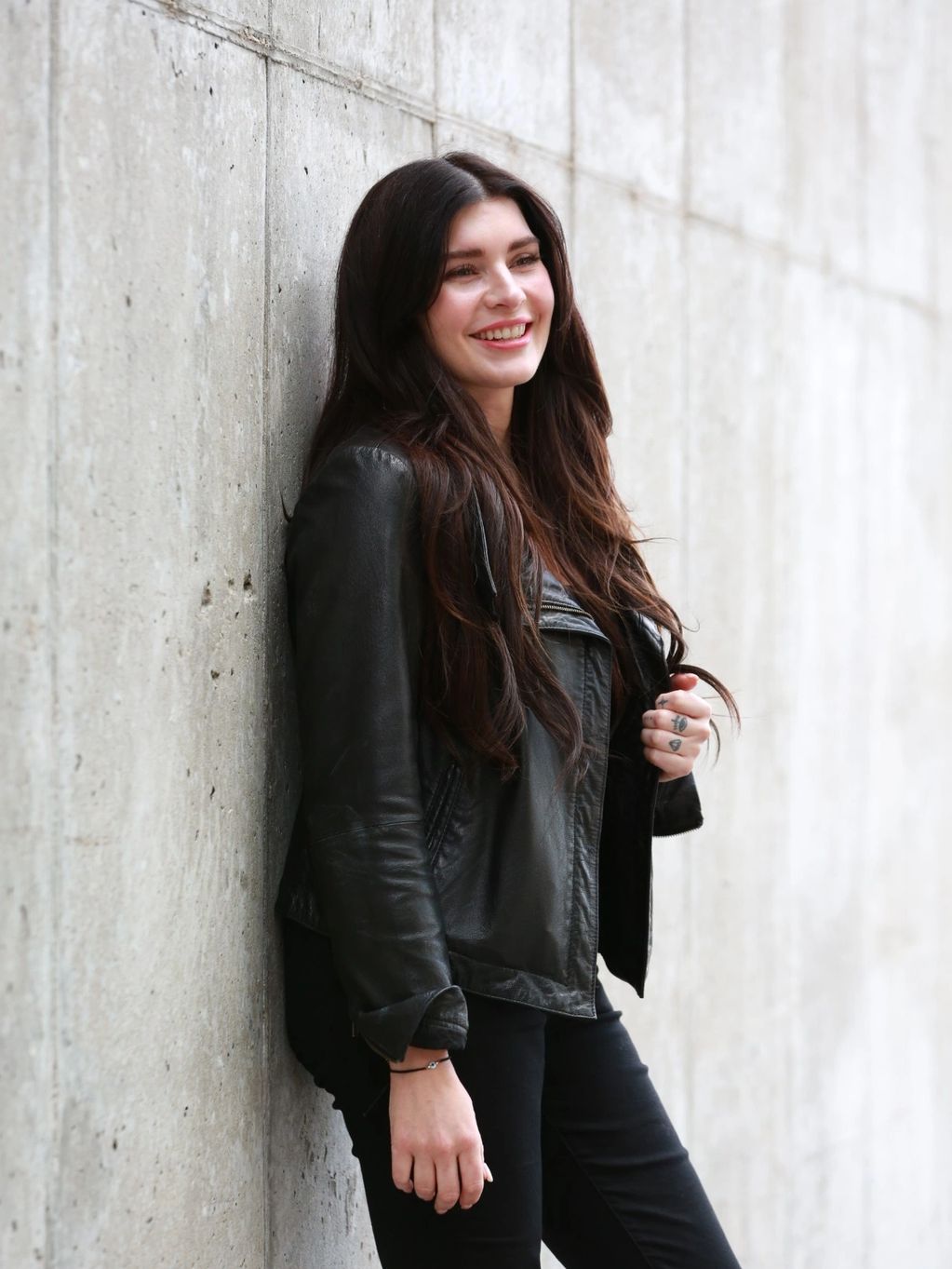 Smiling woman with long dark hair leaning against a concrete wall, wearing a black leather jacket.