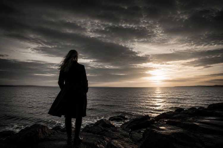 A person standing on rocky shore at sunset.