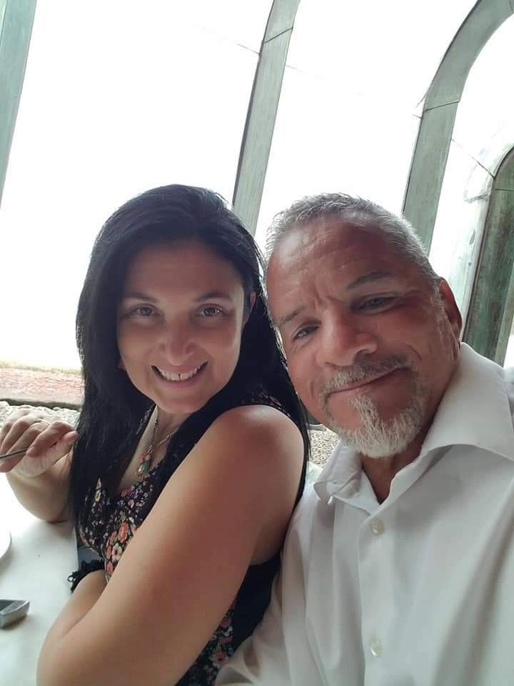 A smiling couple sitting together indoors near large windows.