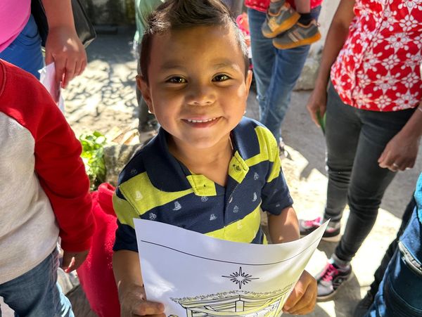 Smiling boy holding a drawing, surrounded by people outdoors.