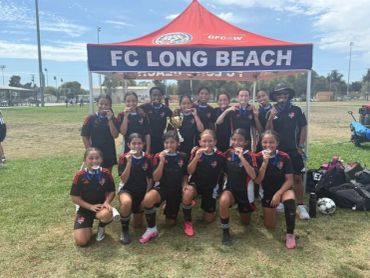 Youth soccer team posing with medals under FC Long Beach tent.