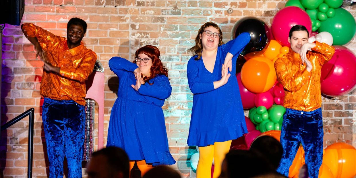 Four people dancing in colorful outfits with vibrant balloon decorations behind them.