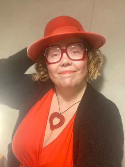Woman in red hat, glasses, and dress with heart necklace.