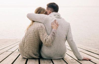 The back of a Couple cuddling sitting on a wooden jetty leading out to water 
