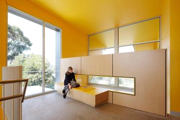 Person sitting on a bench in a modern yellow room with large windows.