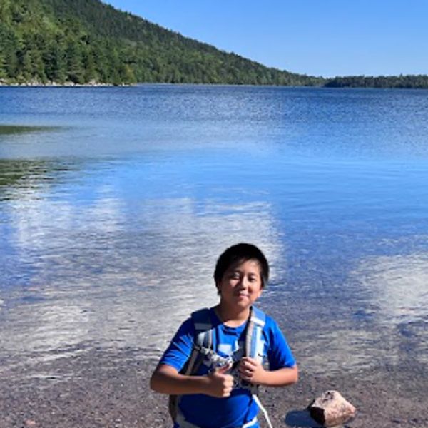 Boy giving thumbs up by a lake with forested hills.
