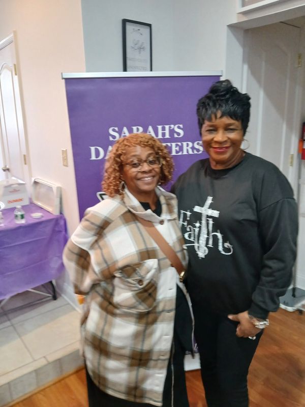 Two smiling women standing in front of a purple banner that reads Sarah's Daughters.