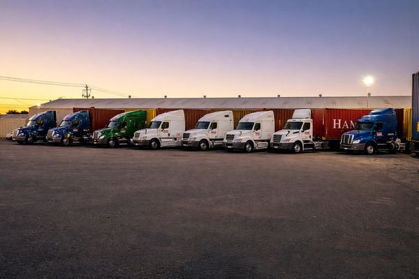 Row of semi trucks parked in front of shipping containers at dusk.