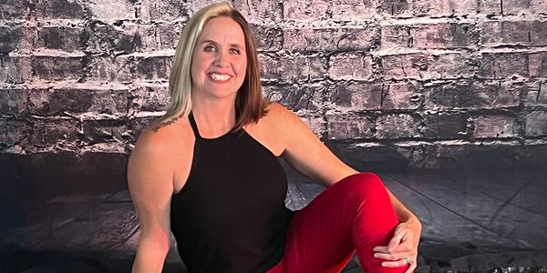 A smiling woman in a black top and red pants sitting against a brick wall.