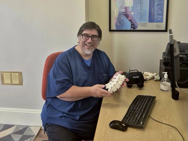 Smiling man in scrubs holding a spine model at desk.