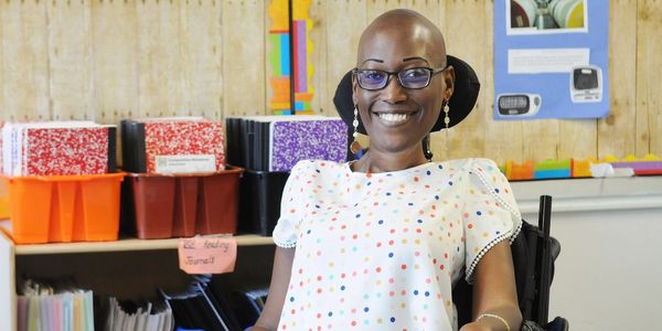 Smiling woman in glasses and polka dot blouse sitting in a wheelchair in an office.