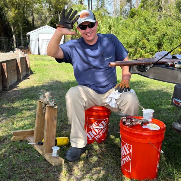 Smiling man in sunglasses waves while sitting on an orange Home Depot bucket outdoors.