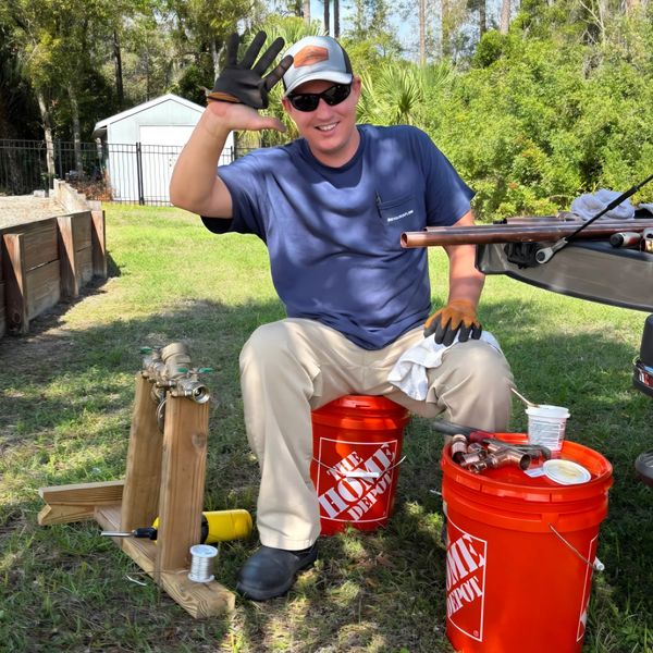 Smiling man in sunglasses waves while sitting on an orange Home Depot bucket outdoors.