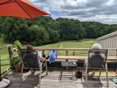 Two people relaxing on chairs on a wooden deck overlooking a green field and forest.