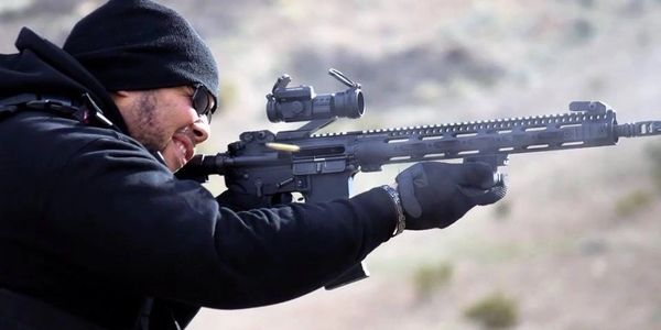 Man in tactical gear firing a rifle outdoors.