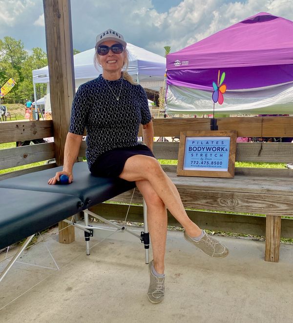 Woman sitting on a Pilates table outdoors, smiling and wearing sunglasses and a cap.