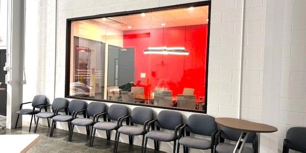 A row of gray chairs lined up against a wall with a window showing a red-walled office.