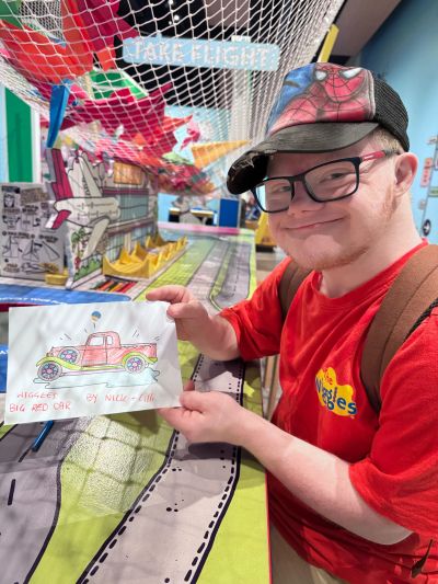 Young man proudly shows his colorful drawing of a red car at a creative art table.