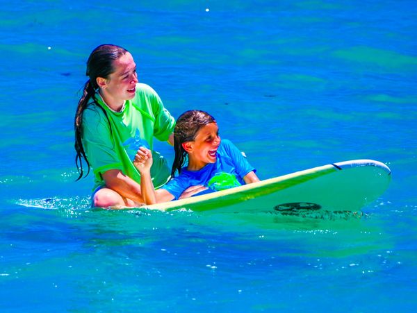 Two girls enjoying a surf lesson on a bright green surfboard in clear blue water.
