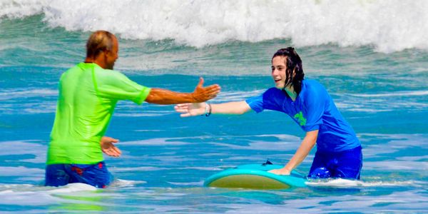 Two people in the ocean reaching out to each other, one with a surfboard.