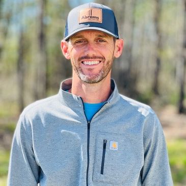 Smiling man wearing a gray jacket and Abrams Construction cap outdoors.