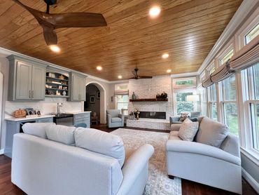 A cozy living room with light gray furniture, large windows, and a wooden ceiling fan.