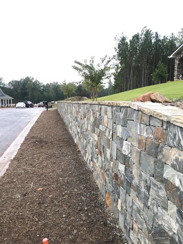 A long stone retaining wall along a residential street with mulch and young trees.