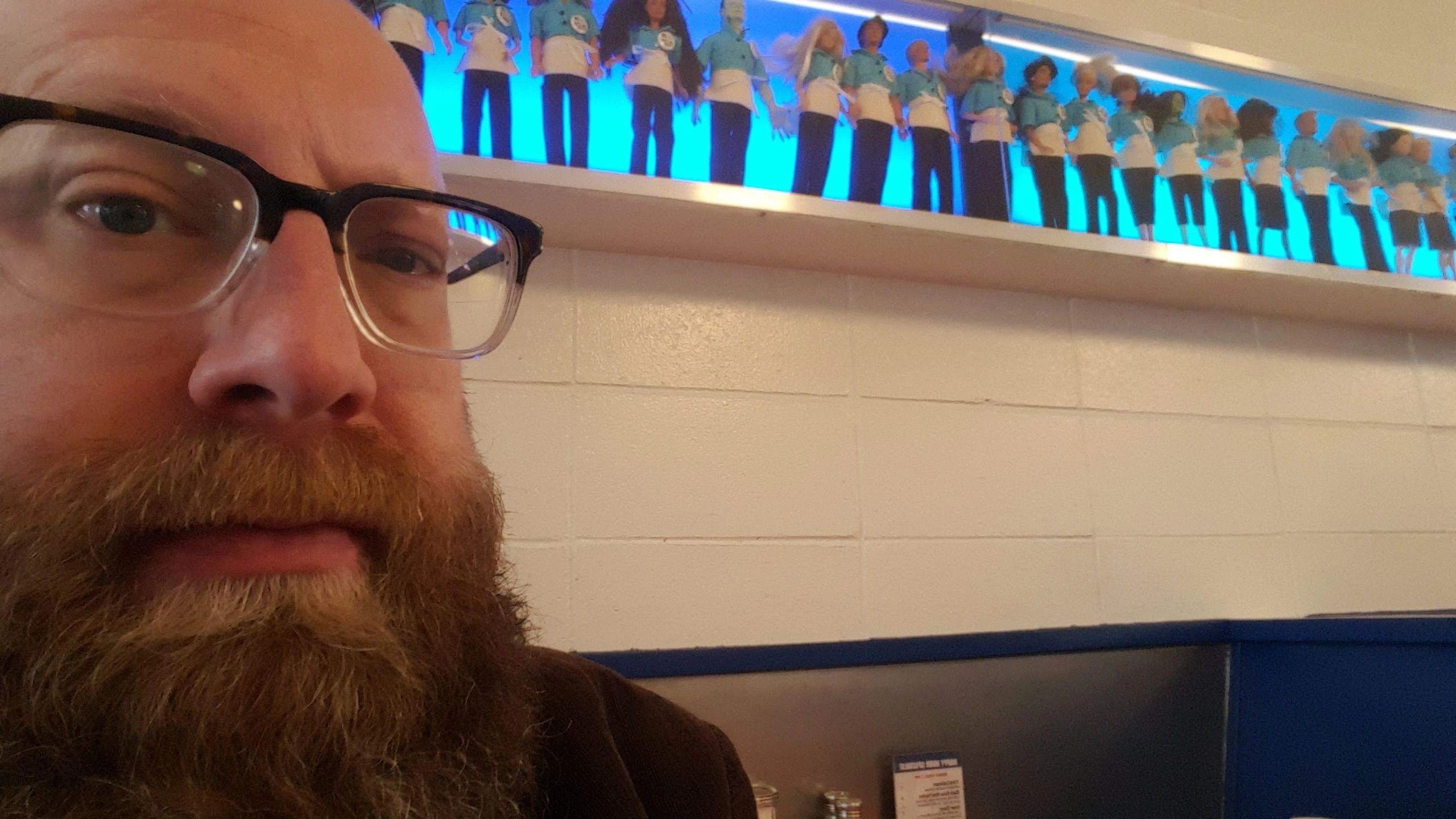 David Leitner in front of a shelf of barbie dolls in blue waiter uniforms backlit with blue light.
