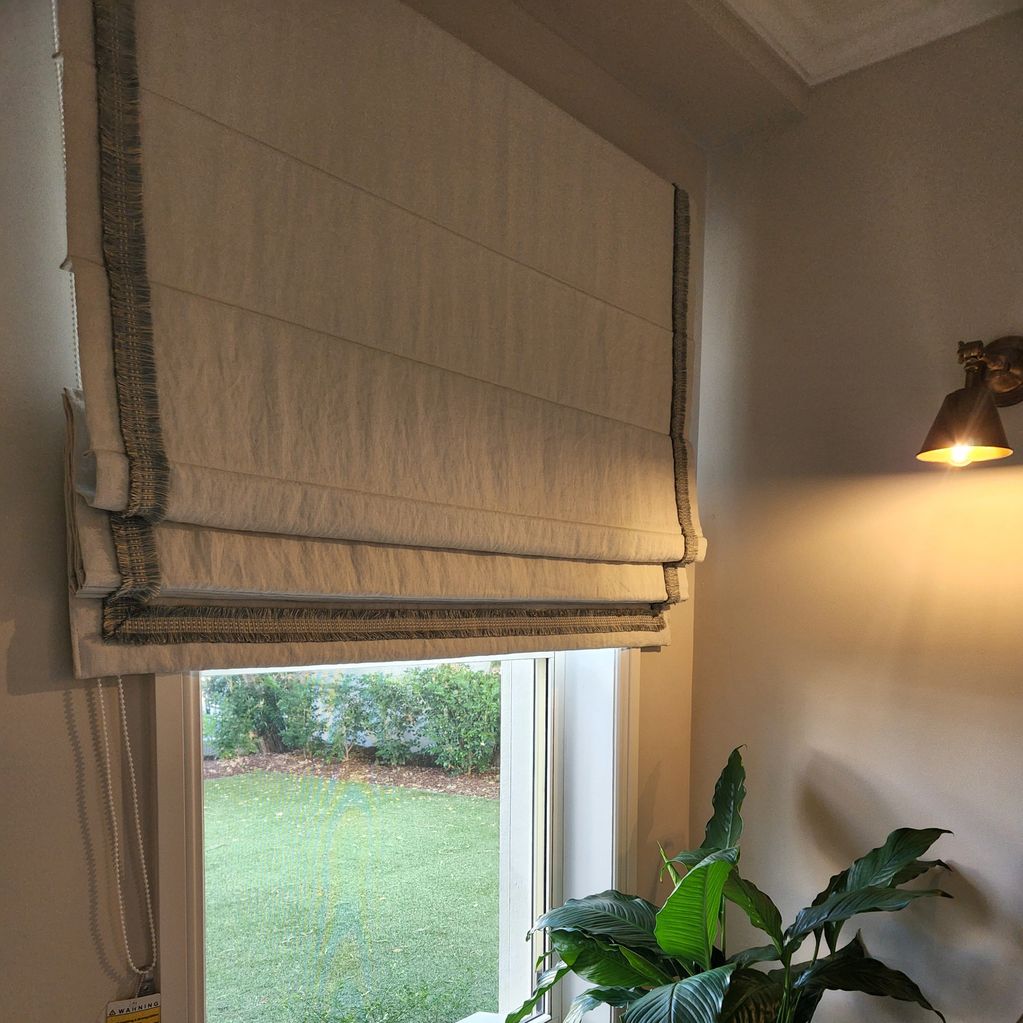 Window with beige roman shade, indoor plant, and wall lamp.