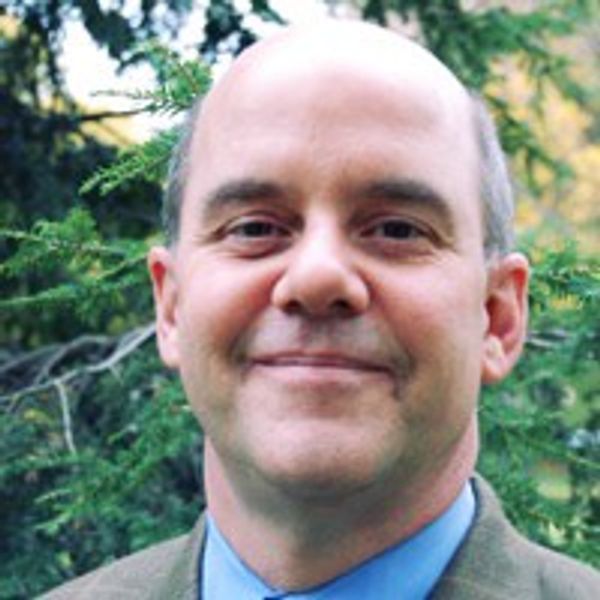 Smiling man in a suit standing outdoors with greenery in the background.