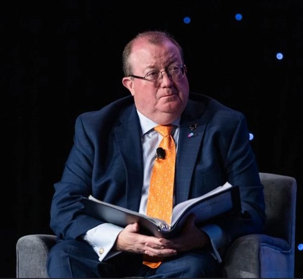 Man in suit and orange tie holding an open book on stage.