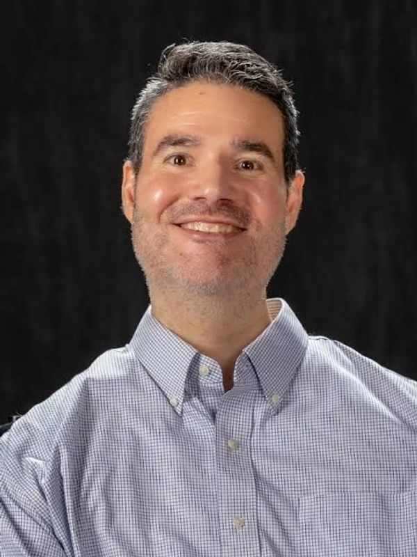 Smiling man in a checkered button-up shirt against a dark background.