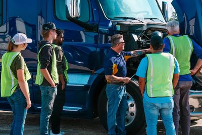 A group of CDL students in safety vests inspecting a blue semi truck's engine.