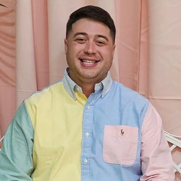 Smiling man wearing a colorful patchwork shirt against a pink curtain backdrop.