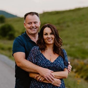 A couple embracing outdoors with smiles and a green hillside in the background.