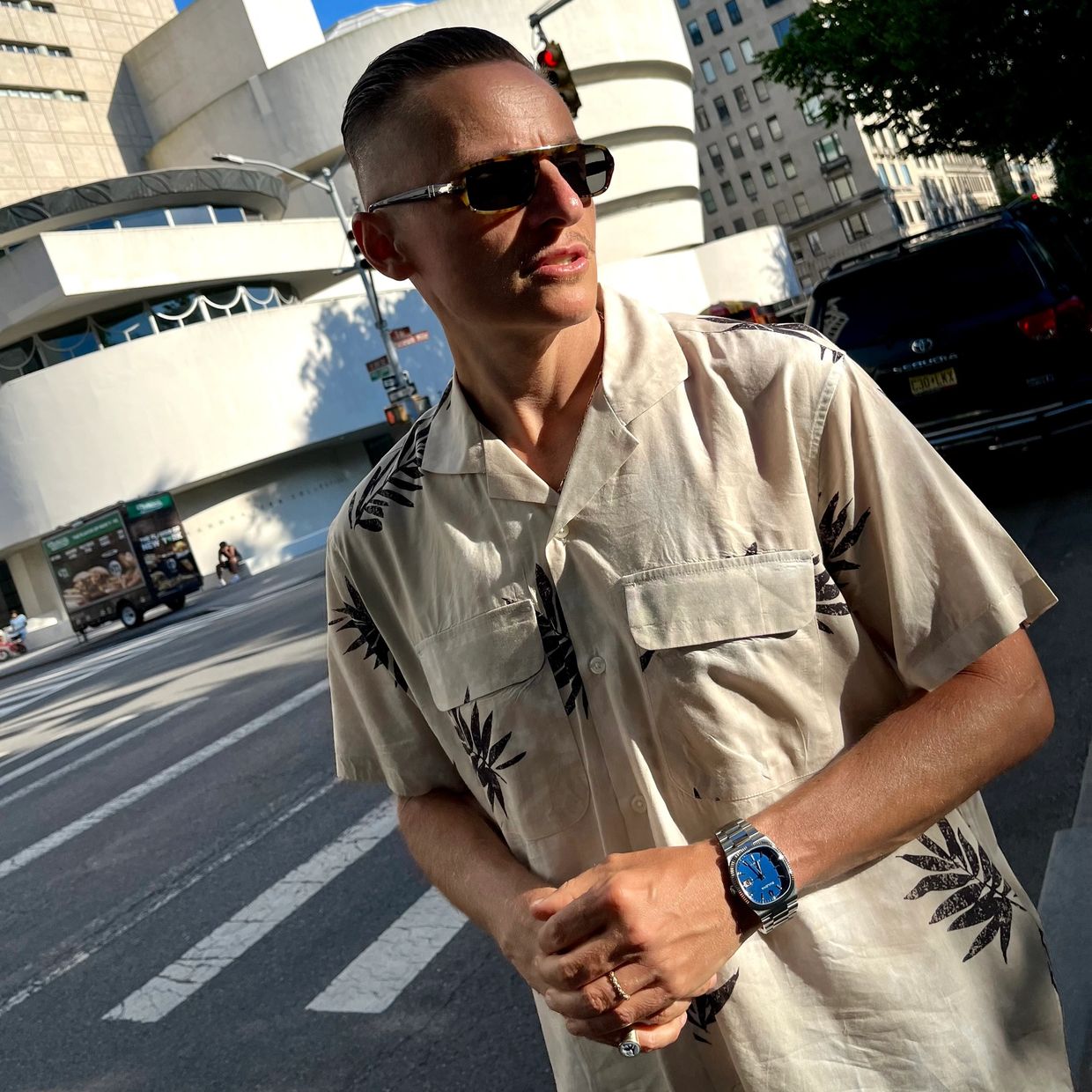 Man in sunglasses and printed shirt standing on city street.