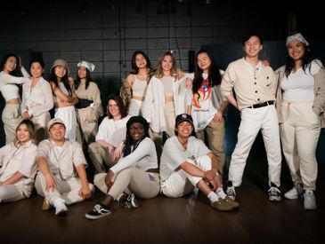 A diverse group of young people posing together in casual light-colored clothing indoors.
