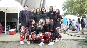 A group of nine young people wearing matching black tops and red camo pants pose at an outdoor event.