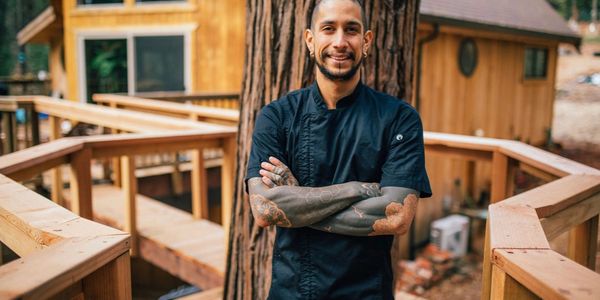 Smiling tattooed man in chef's coat standing outdoors on wooden deck.