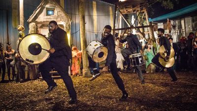 Masked musicians energetically perform with drums and cymbals at a lively nighttime outdoor event.