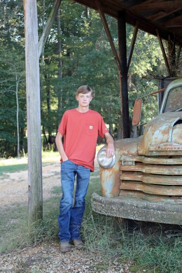 Young man in red shirt leaning on old rusted truck.
